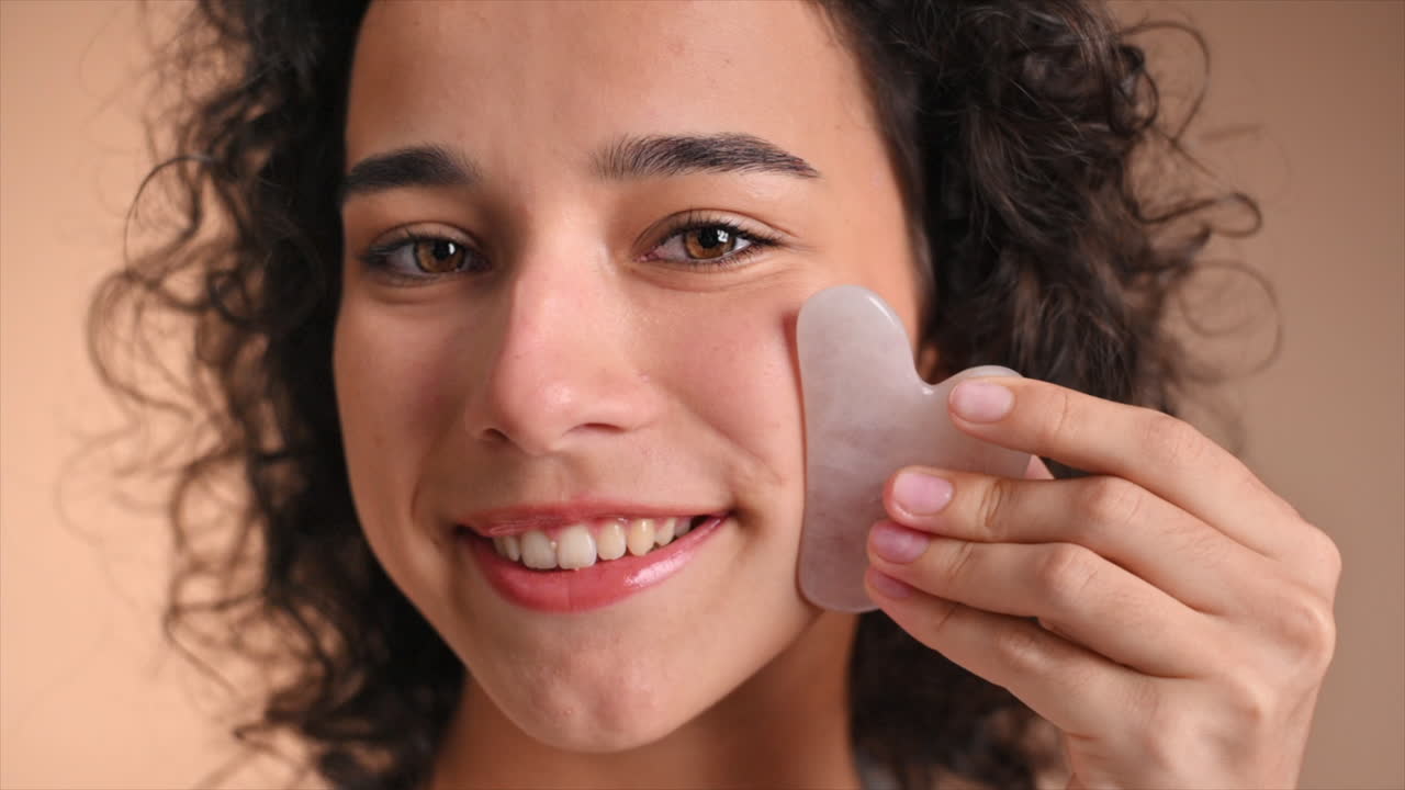 A young caucasian smiling woman is doing a facial massage using Gua Sha, looking into the camera. Slow motion