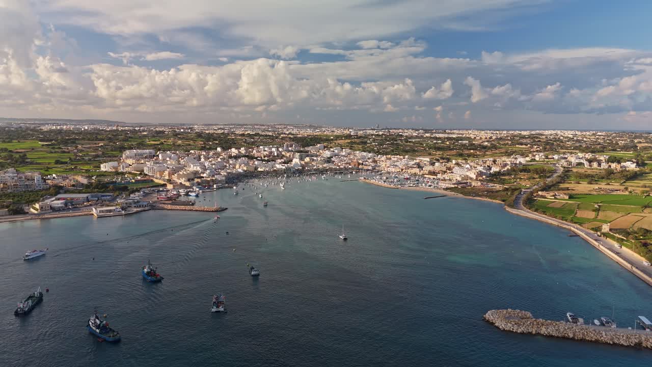 Vista aérea de Marsaxlokk, Malta, mostrando seu pitoresco porto com barcos de pesca coloridos, a costa mediterrânea e a zona rural circundante sob um céu de inverno parcialmente nublado