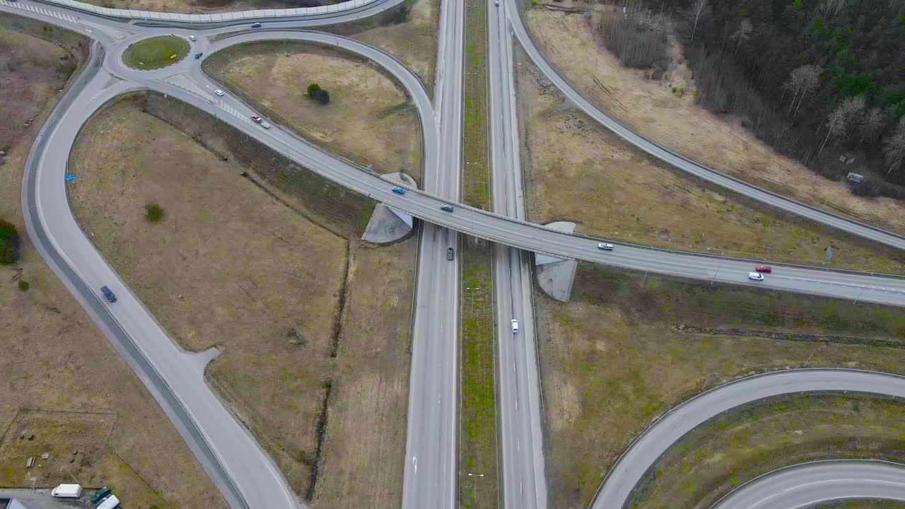 Aerial view of a bridge going over a highway in Estonia that is called Tallinn Pärnu road 2+2. Cars are driving over and under a bridge road and a large intersection is visible in Saue at cloudy day.