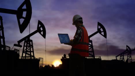 vista trasera de un ingeniero masculino asiático con casco de seguridad mirando el plano de la turbina eólica en su portátil mientras está de pie frente a las bombas de petróleo, durante la puesta o el amanecer