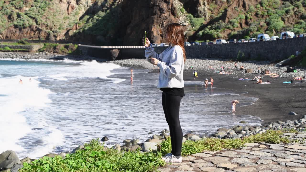 una joven de cabello castaño toma una foto de paisaje en una playa de arena negra con su teléfono móvil