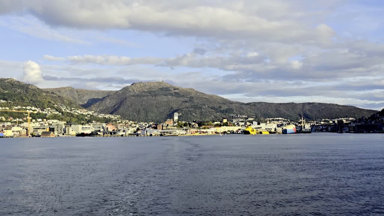 Distant view from aft deck as vessel departs Bergen harbor with skyline and fjord in evening light