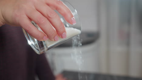 close-up of a person hand holding a glass cup filled with sugar, gently pouring it in a modern kitchen, the sugar flows smoothly, creating a dynamic visual of food preparation and baking