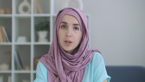 A-Muslim-woman-in-a-hijab-sits-at-a-table-and-looks-into-the-camera-waving-her-hand-and-talking.-vlog-or-video-call-a-woman-in-a-hijab.-shot-of-the-head-of-an-Arab-woman-in-conversation