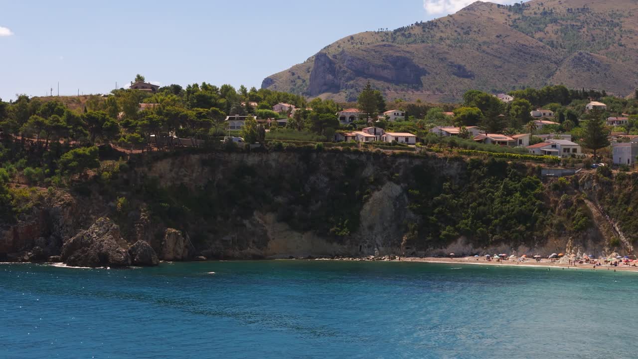 Panning left aerial drone shot of coastal cliff at Guidaloca Beach (Spiaggia di Guidaloca) in Scopello, Sicily, Italy, during sunny Mediterranean summer travel