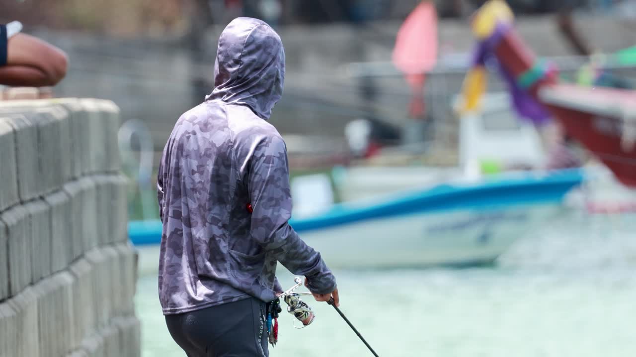 A man fishes near colorful boats in Phuket, Thailand, under bright daylight, capturing a serene coastal scene