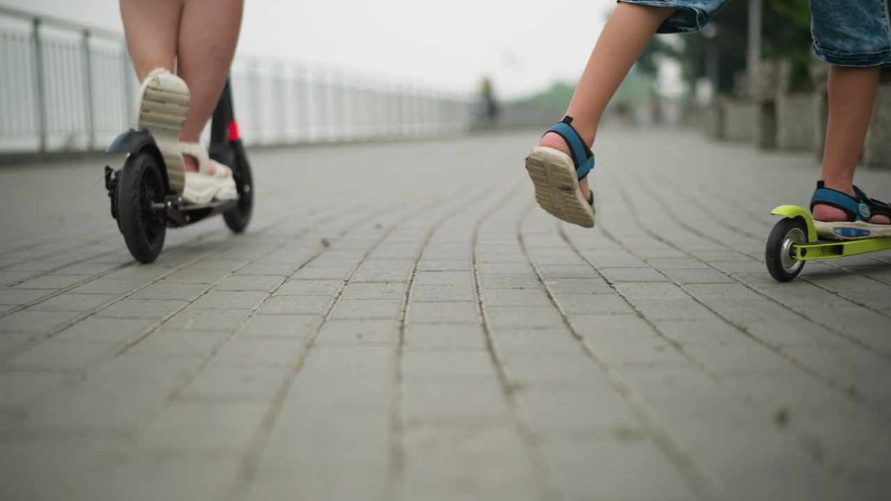 Close-up of a child and a woman riding scooters side by side, the child rides a green scooter, while the woman is on a black one