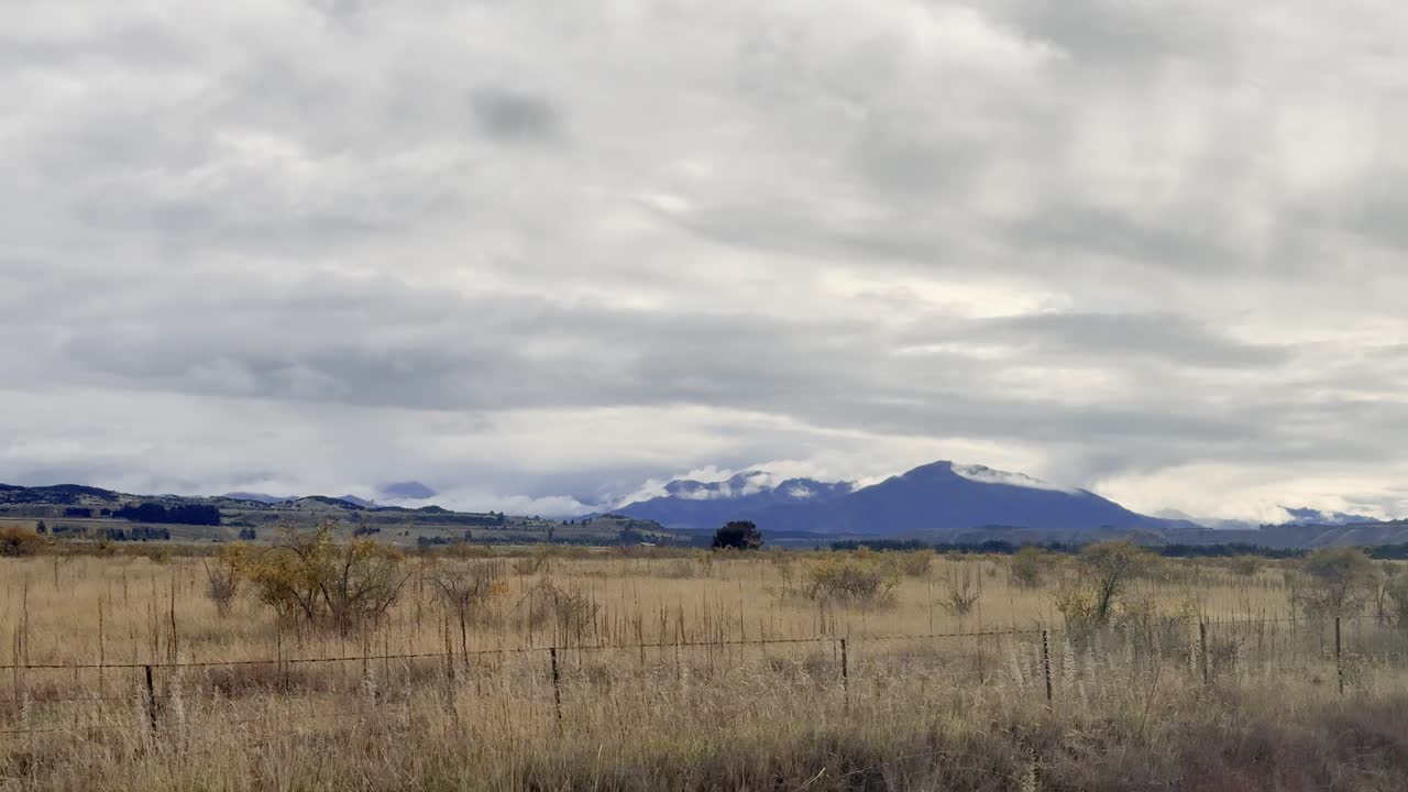 A moving vehicle captures overcast rural farmland with wire fencing, dry grass, and distant snow-capped mountains near Wanaka, New Zealand. Subtle camera panning, natural light
