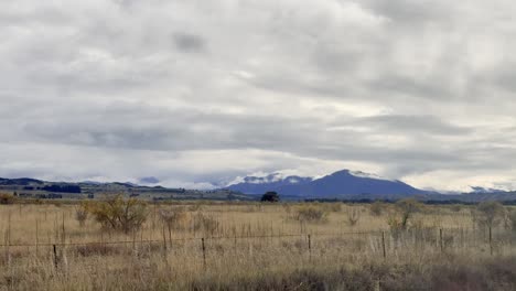 a moving vehicle captures overcast rural farmland with wire fencing, dry grass, and distant snow-capped mountains near wanaka, new zealand. subtle camera panning, natural light