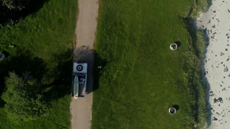 Top-view-of-a-guy-driving-in-his-land-rover-with-a-surf-board-on-a-paved-road-between-green-patches-of-grass-next-to-the-beach