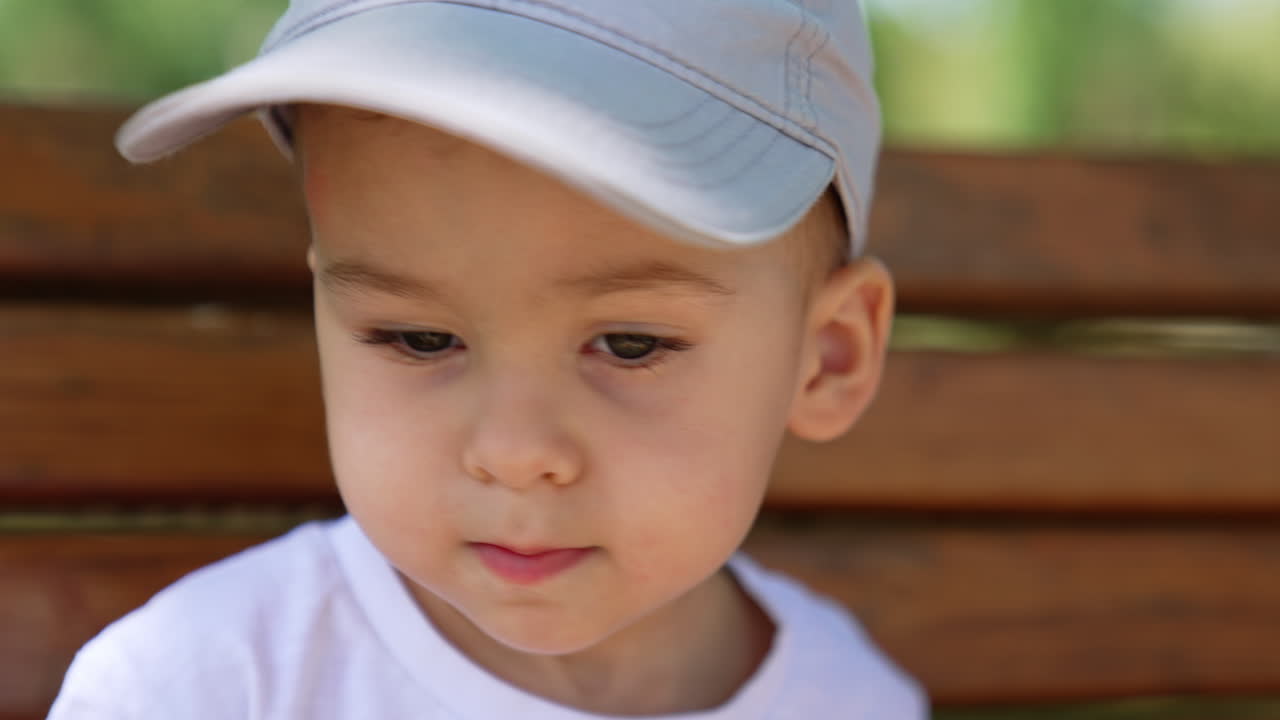 Adorable sad baby boy's face in cap sitting outdoors. Calm kid waving head negatively. Close up.