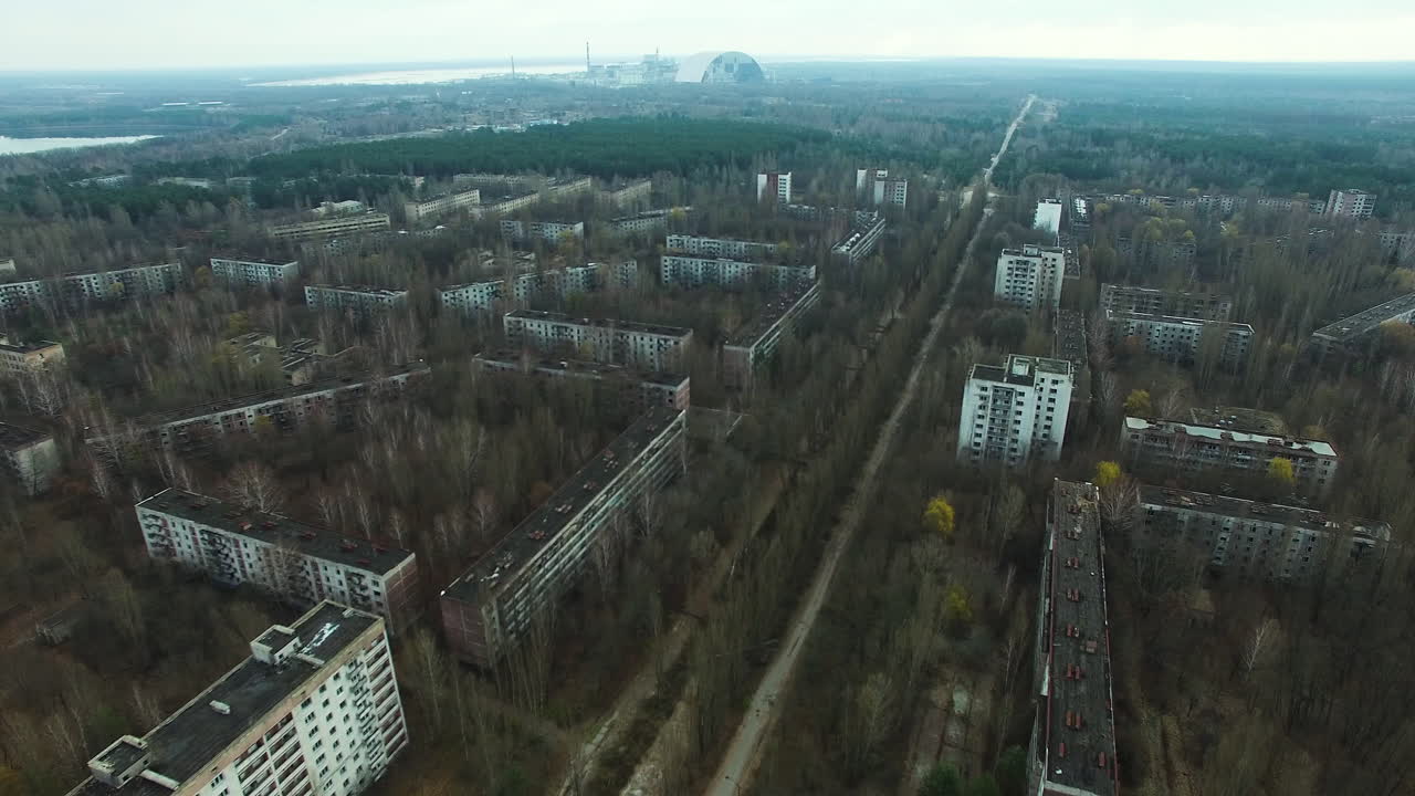 Кадры с дрона Припяти, Украина, заброшенного города рядом с Чернобыльской атомной электростанцией. Жилые кварталы и дороги заросли густыми лесами. Вдалеке виднеется объект «Новый безопасный конфайнмент» Чернобыльской АЭС.