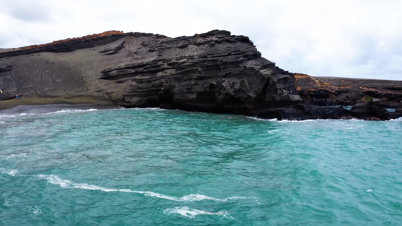 Vivid turquoise sea gently laps against rugged volcanic cliffs and a black sand shoreline, showcasing the raw beauty of this unique coastal landscape.