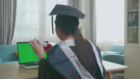 excited asian woman showing off a university certificate to laptop computer with green mock-up screen during an online video call. pretty female graduate wearing a graduation gown and cap sitting on the living room
