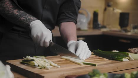 Fresh-zucchini-being-sliced-on-a-wooden-board-by-young-professional-male-chef-in-an-elegant-black-shirt-with-tattoos