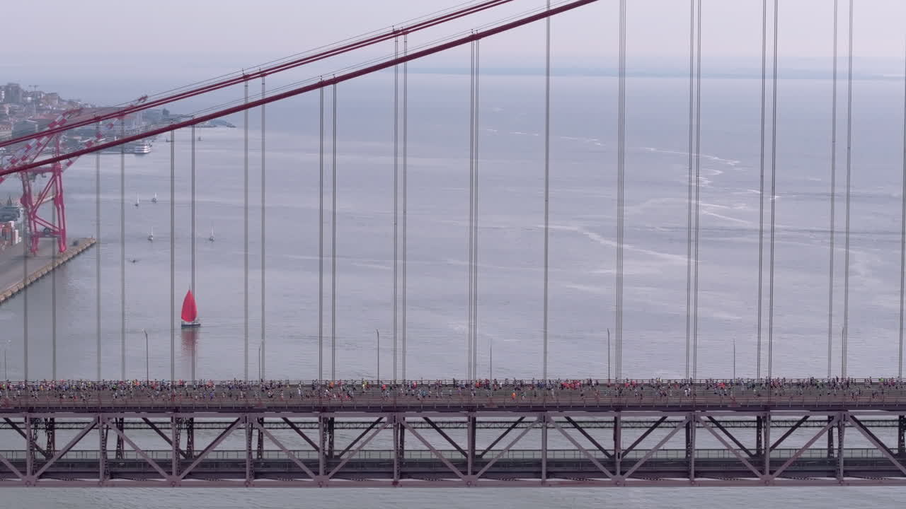 Side-on aerial drone shot of half marathon and 10K long-distance running event in Lisbon, Portugal, Europe. Runners crossing the iconic famous red 25th April suspension bridge