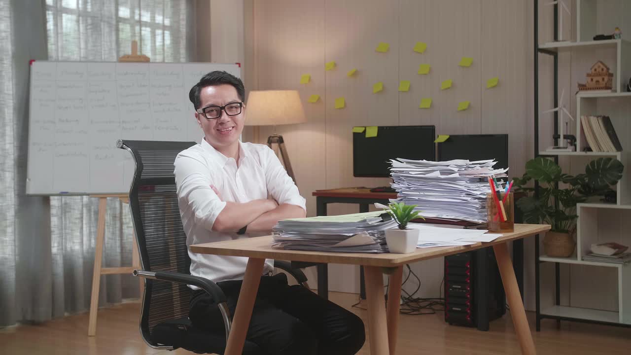 hombre asiático cruzando los brazos y sonriendo a la cámara mientras trabaja con documentos en la oficina