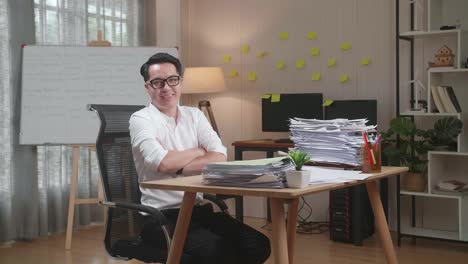 hombre asiático cruzando los brazos y sonriendo a la cámara mientras trabaja con documentos en la oficina