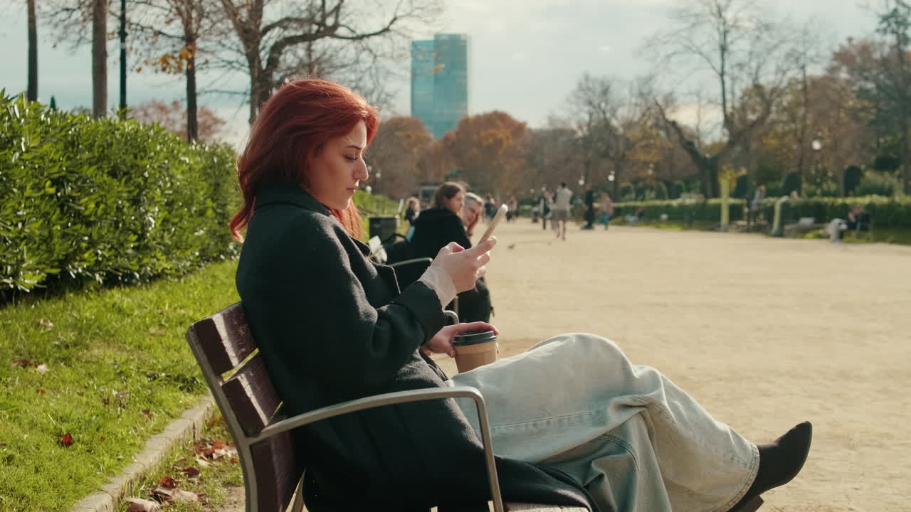 Ung rødhåret kvinde scroller på smartphone i parken med kaffe