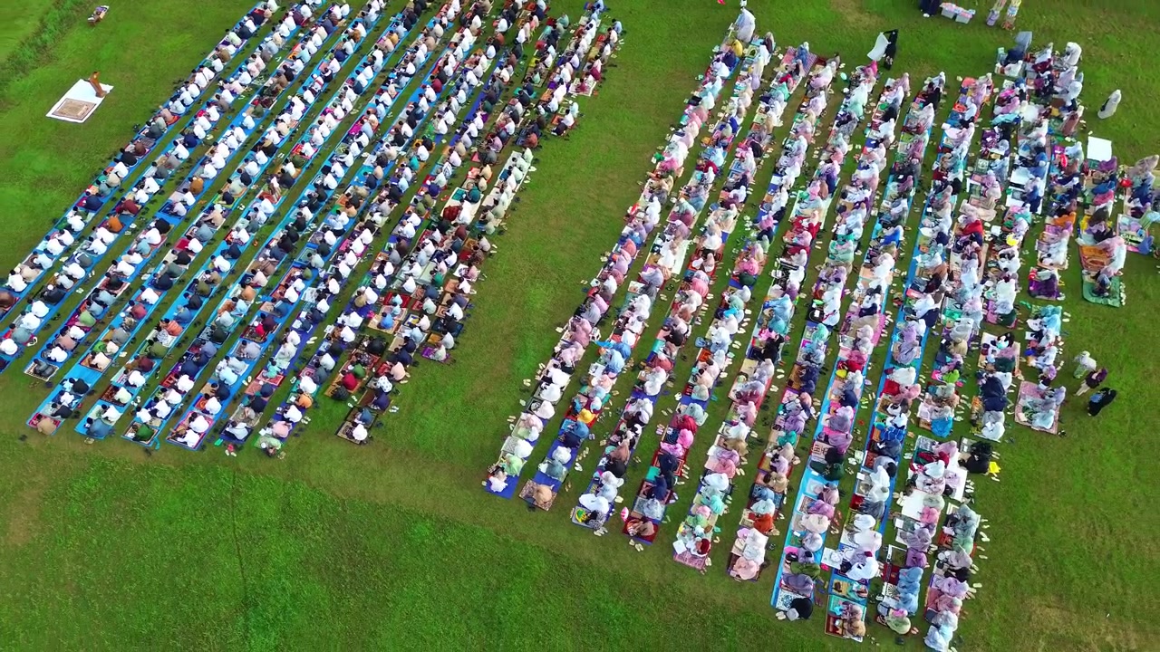 Drone shot of Muslim congregation praying Eid prayer outdoors, showcasing unity and faith on green field.