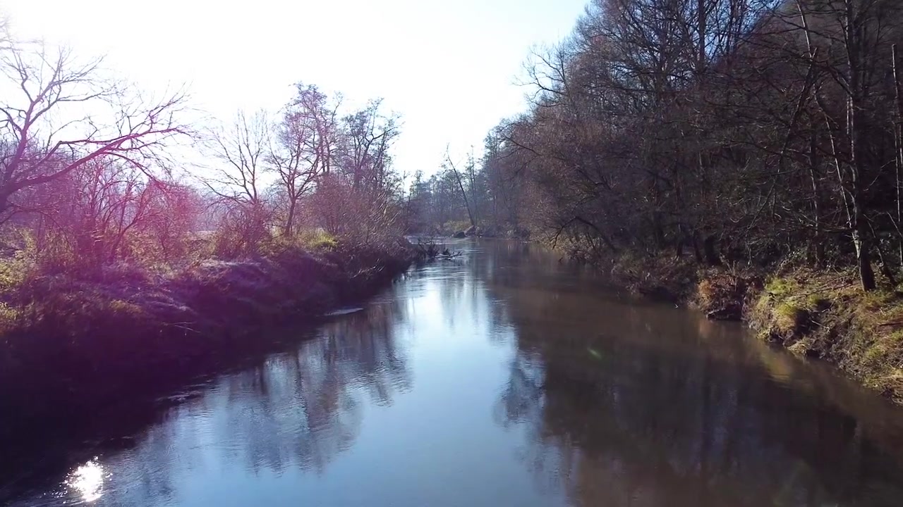En rolig udsigt over en flod omgivet af nøgne træer i det sene efterår eller den tidlige vinter. Det stille vand reflekterer den klare sol og det omkringliggende landskab, hvilket indfanger naturens fredfyldthed
