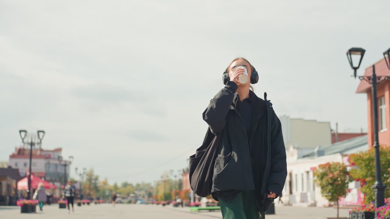 confident woman walking with coffee and shoulder bag, steady stride through open plaza, urban architecture and lampposts in frame, purposeful routine with relaxed vibe and cinematic composition