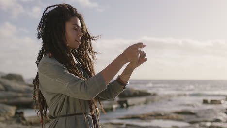 hermosa mujer de raza mixta con rastas tomando fotos en la playa usando el teléfono sonriendo feliz