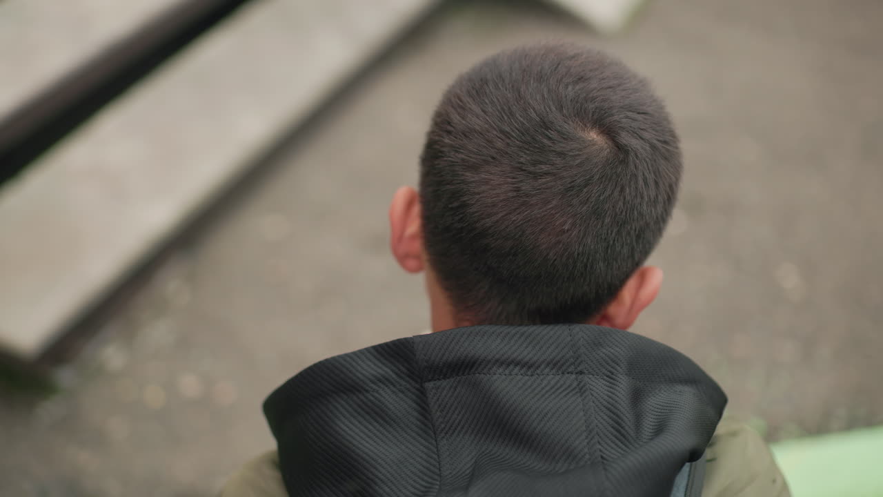 Close up overhead view of boy wearing green jacket and backpack concentrating on study while reading book outdoors with blurred ground and stair background