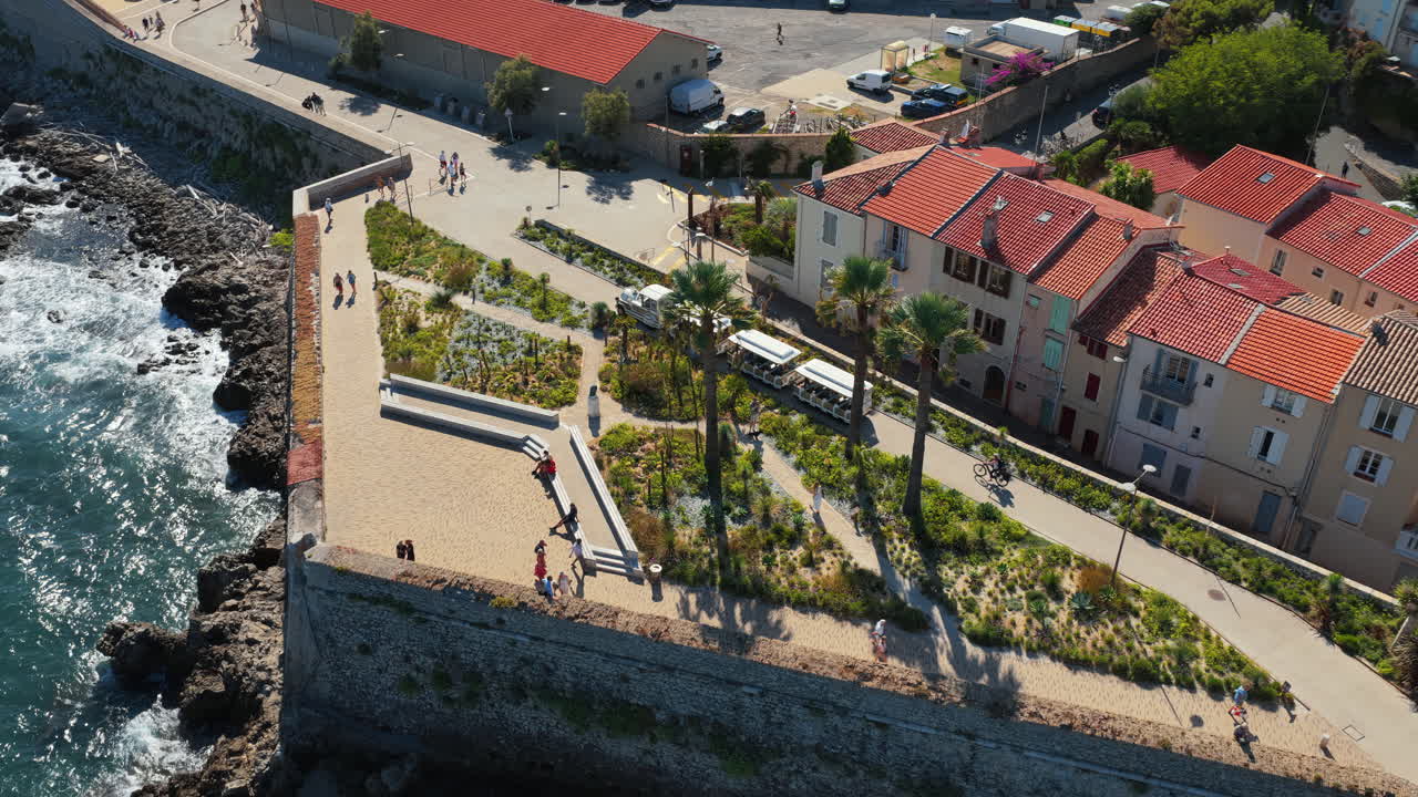 Aerial drone view of people walking along the Antibes Old Town ramparts, with palm trees, landscaped gardens, and waves crashing against the rocky coast