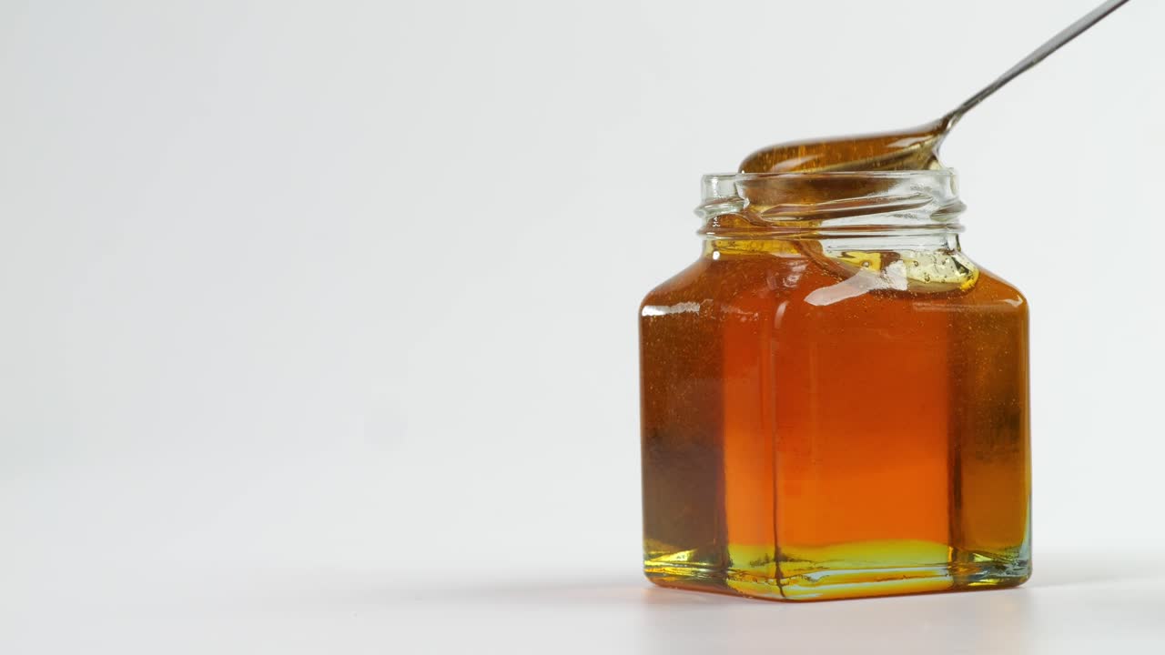 A Metal Spoon Is Dipped To Take Honey In A Jar On The White Background. Honey Dripping From Metal Spoon, Healthy Organic Thick Honey, Sweet Honey Dessert 4K. Slow Motion