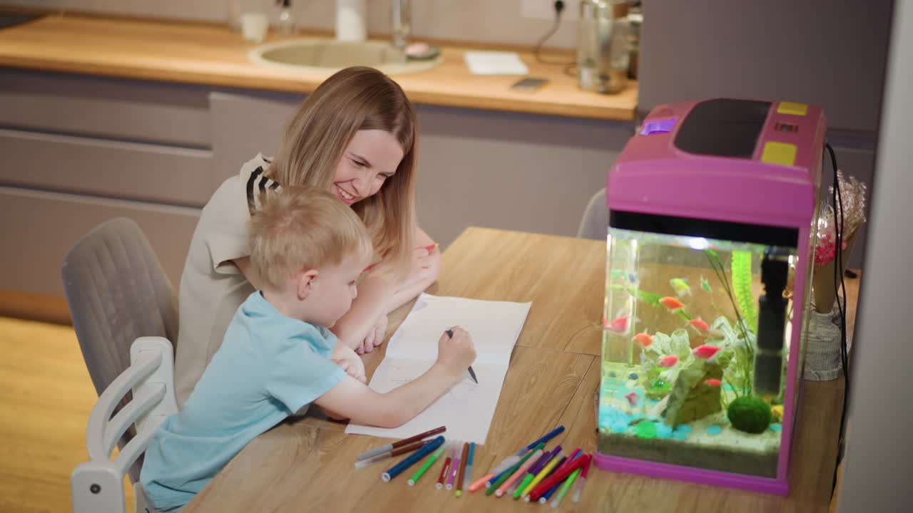 Mother and young boy drawing together with colorful markers on paper at wooden table beside bright aquarium filled with vibrant fish, enjoying creative activity and bonding time in cozy home environment