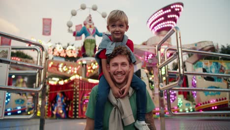 portrait of a happy father with curly hair in a green t-shirt and his little blond son in a red t-shirt against the backdrop of bright lights and attractions in an amusement park