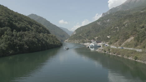 Drohnenschuss-Beim-Flug-über-Die-Fähranlegestelle-In-Fierze,-Albanien,-Mit-Einem-Vorbeifahrenden-Boot-Und-Einer-Fähranlegestelle-Im-Wasser-An-Einem-Sonnigen,-Bewölkten-Tagesprotokoll