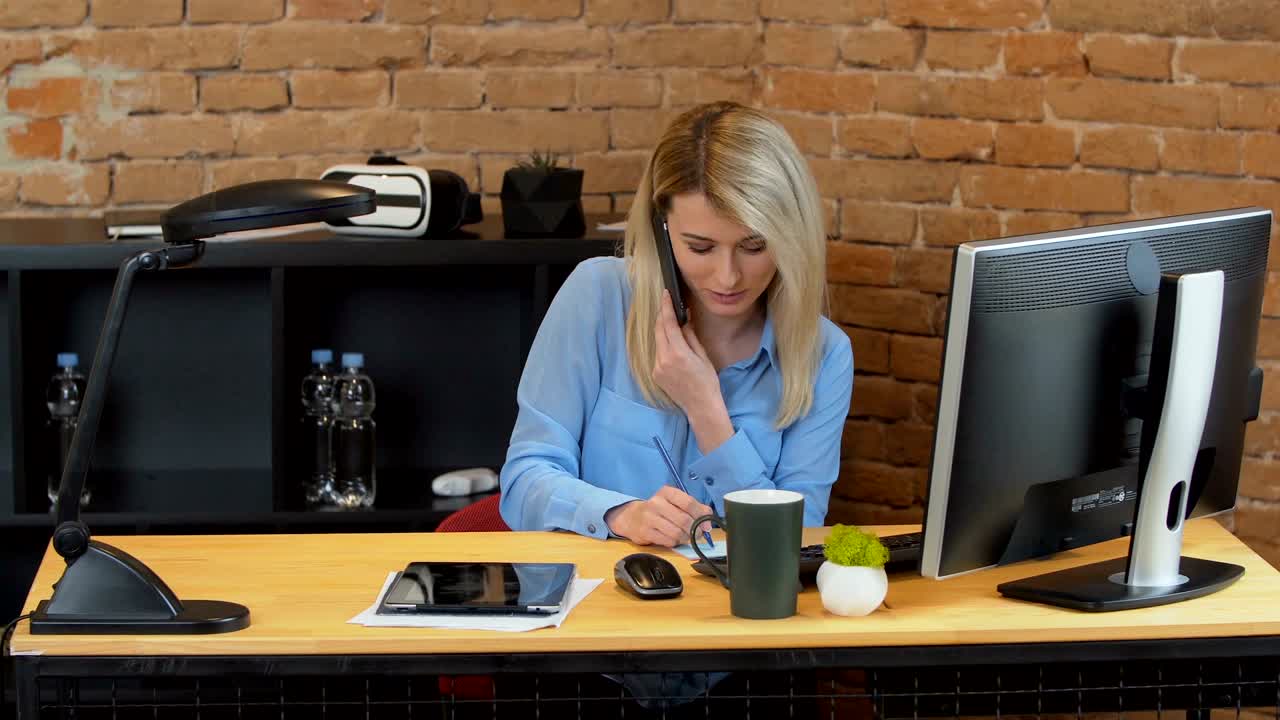 Smuk kvindelig kontoransat besvarer et telefonopkald ved sit skrivebord på et travlt kontor