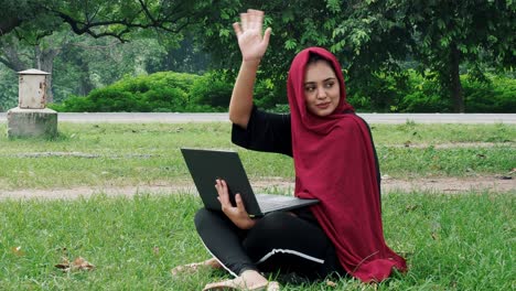 young afghan female student sits on the ground in a park on a sunny day, studies using a computer and does homework and waves hand