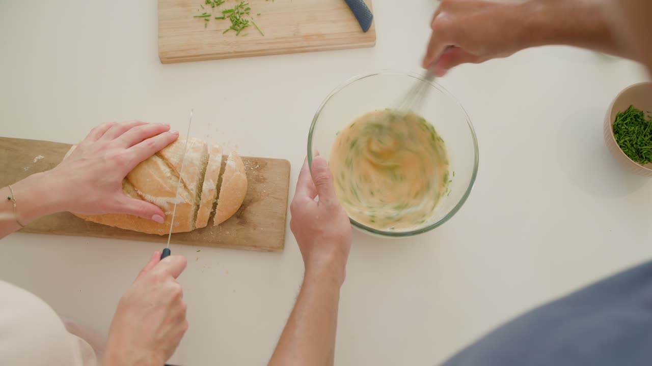 Donna che inizia ad affettare una pagnotta su un tagliere mentre l'uomo sbatte una pastella alle erbe in una ciotola di vetro, cucinando.