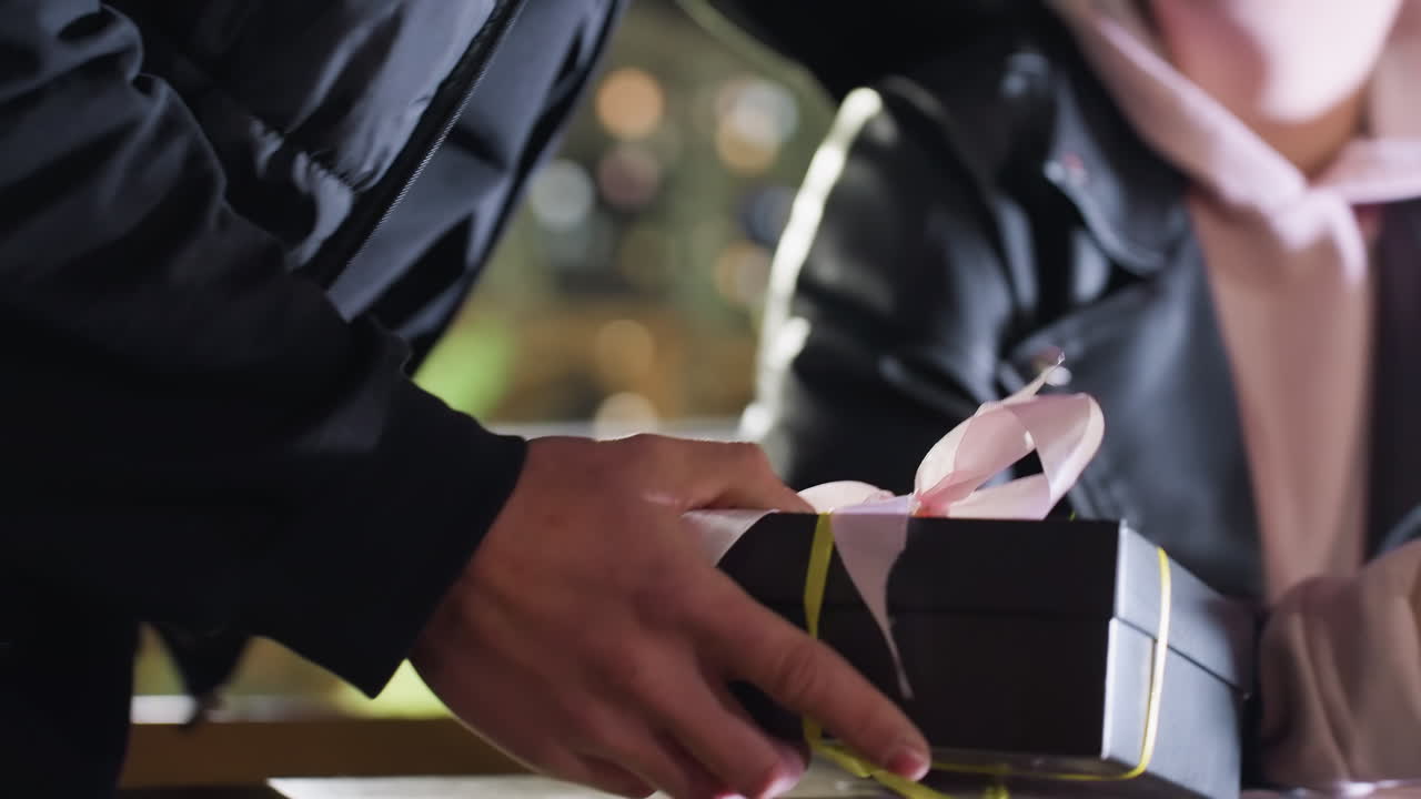 Close up of man holding wrapped gift box with ribbon as he presents it to woman reading red cover book at wooden table under glowing city lights with warm atmosphere and bokeh in background