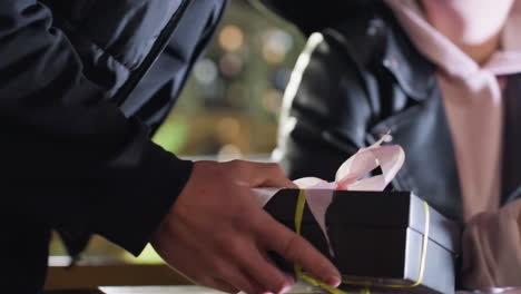 close up of man holding wrapped gift box with ribbon as he presents it to woman reading red cover book at wooden table under glowing city lights with warm atmosphere and bokeh in background