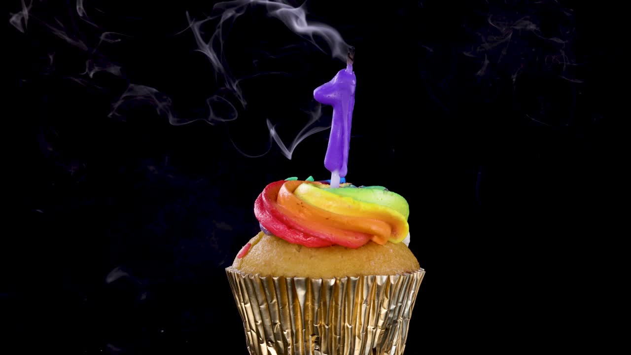 Un singolo cupcake con glassa arcobaleno e una candela accesa a forma di numero uno viene spenta, lasciando scie di fumo su uno sfondo nero in un'inquadratura statica.