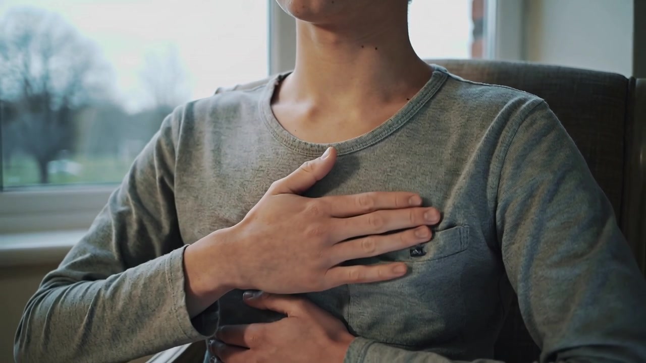 Placing hand on chest and throat, teen holding abdomen, sitting by window wearing grey shirt