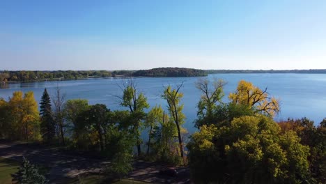 aerial of a drone flying over trees in waseca minnesota during fall and revealing a lake and forest in the background with blue skies