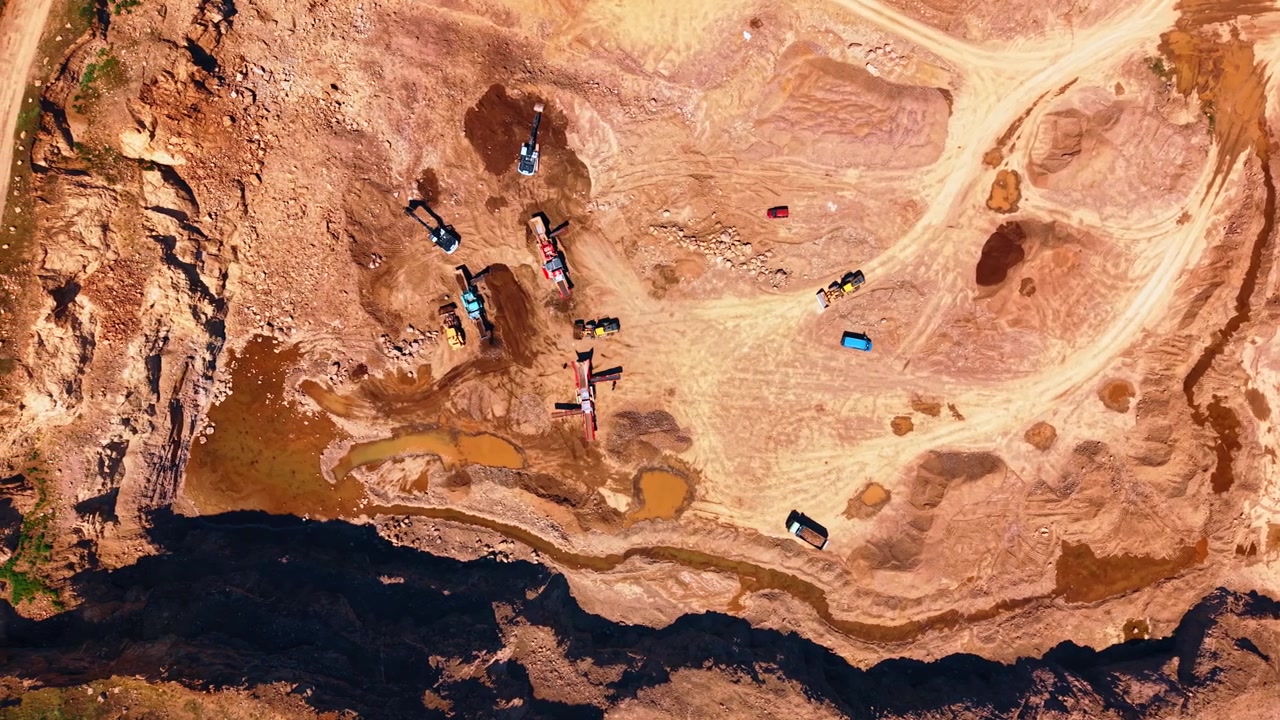 Aerial view of industrial stone quarry and dirt roads. Construction machinery and transport vehicles work on dry brown soil near deep shadow.