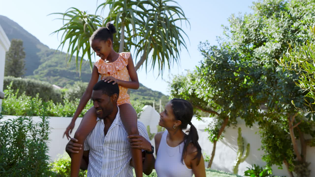 African american father carrying daughter on shoulders, mother walking beside, enjoying sunny garden