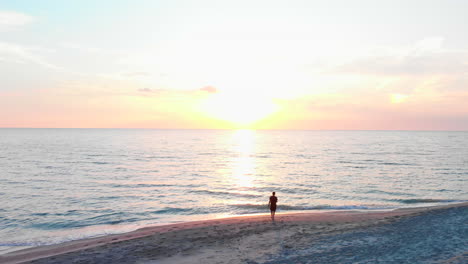 Aerial,-adult-male-standing-on-beach-watches-sunrise,-slow-motion-flyover