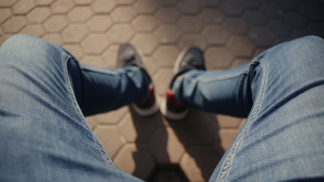 屋外の舗道に座っている男性の、ジーンズとスニーカーを履いた脚の俯瞰図。