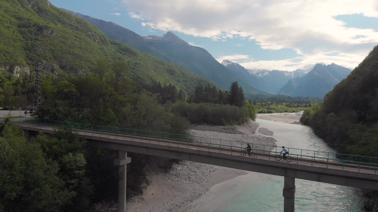 vista lateral aérea de dos ciclistas cruzando el puente a través del río alpino claro, verano