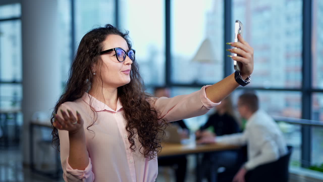 Smiling long-haired woman taking stream video on her smartphone. Female in office using phone for personal needs. People in blur at the backdrop.