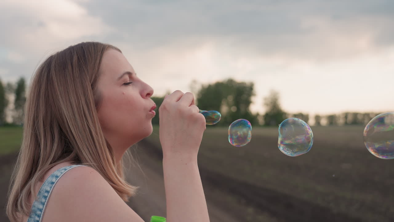 close up side view lady blowing bubbles over expansive farmland at sunset with bubbles drifting under pastel sky capturing serene playful moment of outdoor leisure amid rural landscape