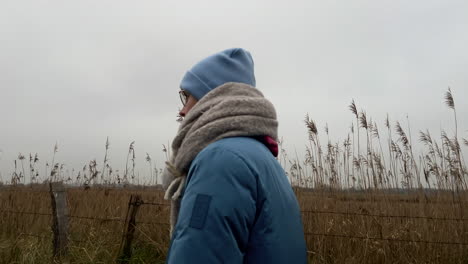 tracking shot of beautiful young woman with scarf and beanie walking between agricultural fields outdoors during cloudy day