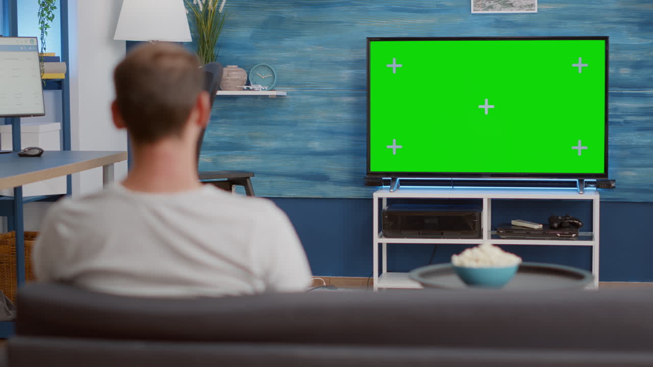 Man sitting on sofa looking at green screen on tv relaxing watching movie in modern living room